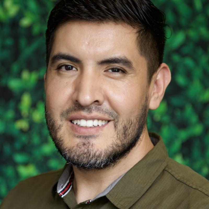 A photo of Enrique Campos wearing an earth green button down shirt with a green background.